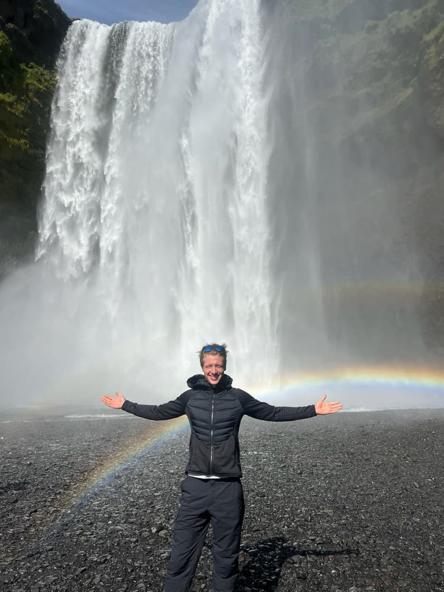 Connor standing with arms outstretched in front of Skógafoss in Iceland, a rainbow arcing across the spray