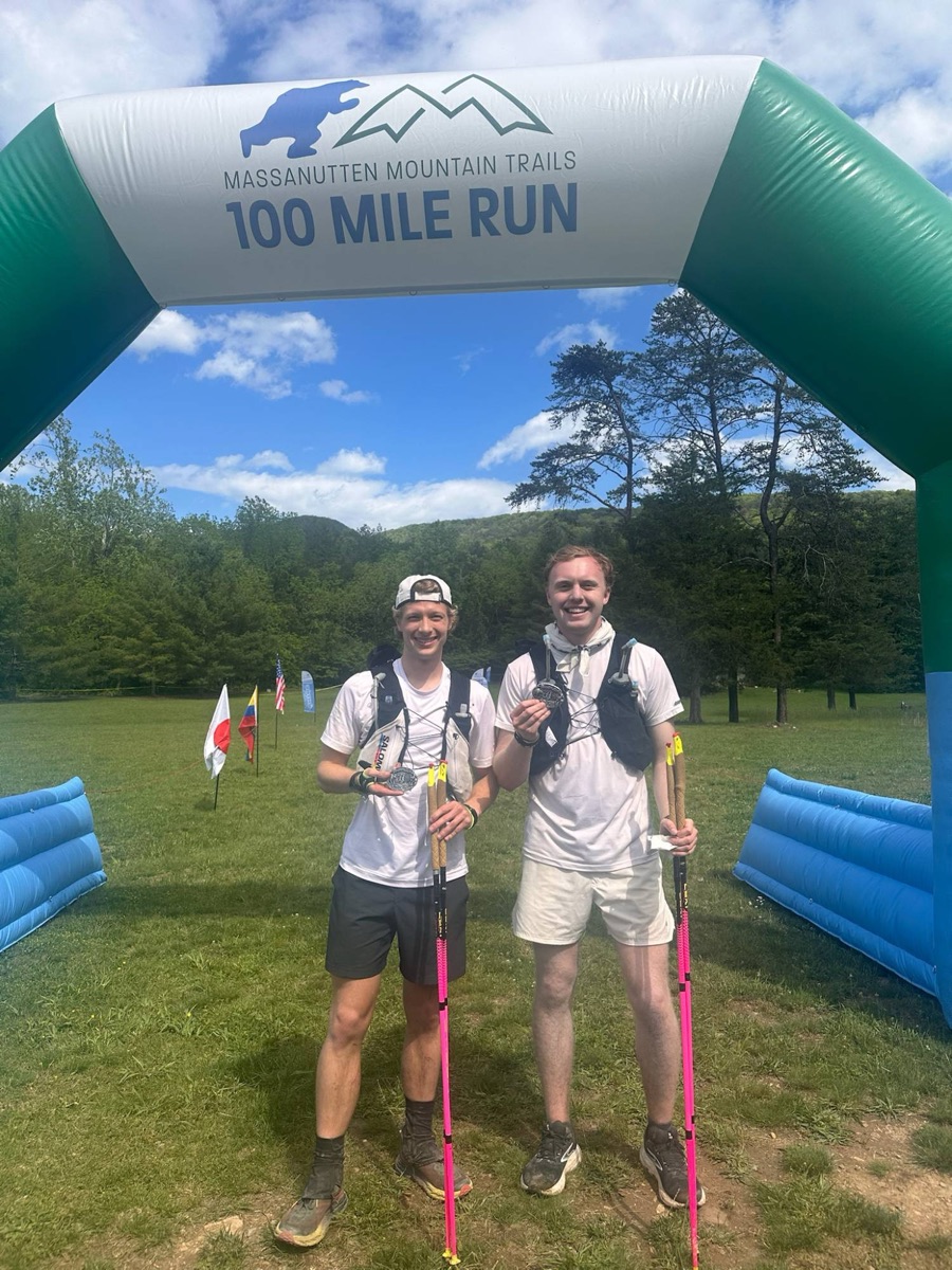 Two runners under the Massanutten Mountain Trails 100 Mile Run finish arch, holding belt buckles and trekking poles